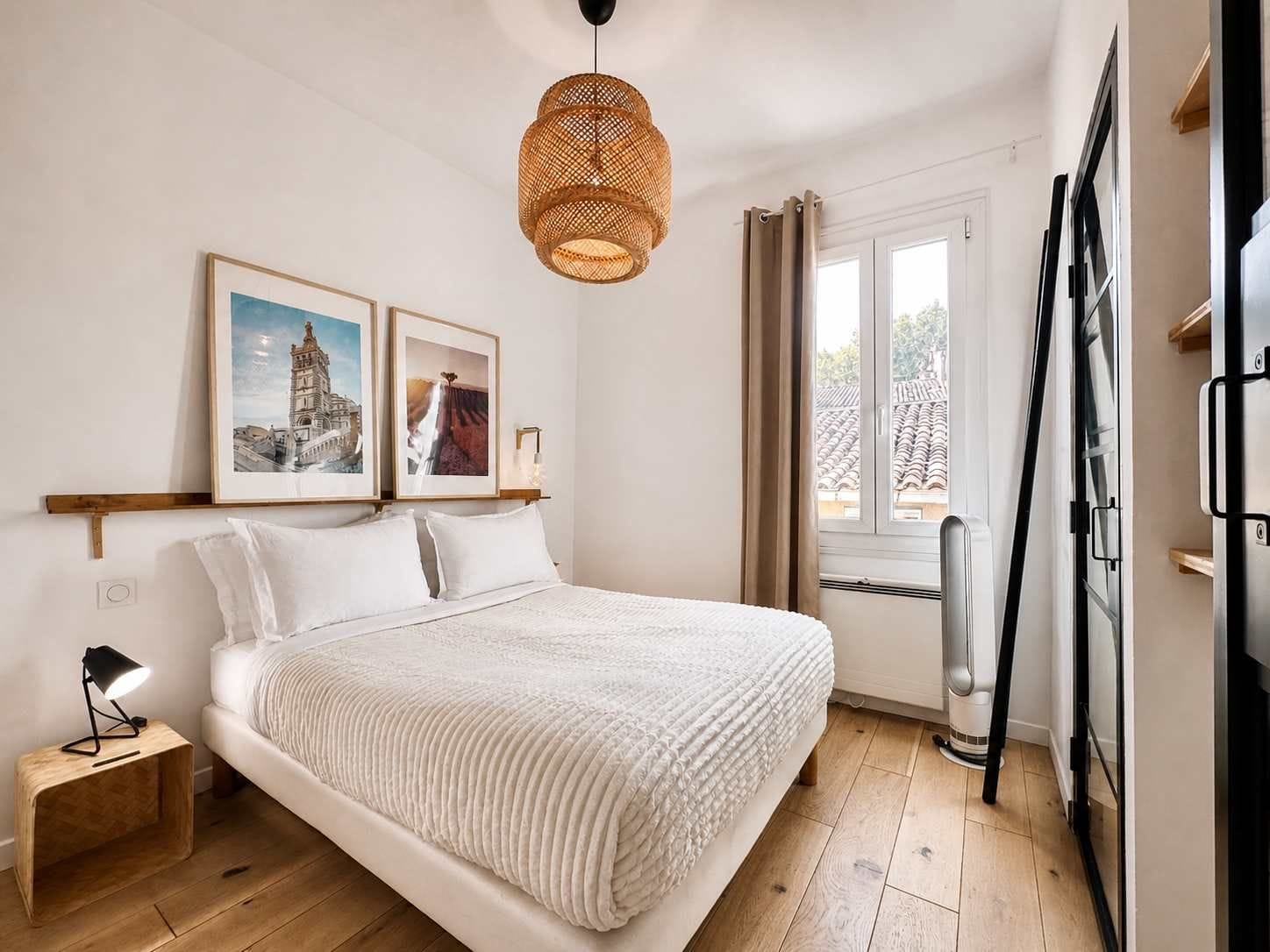 Une chambre à coucher avec un lit, une table de chevet avec une lampe, un luminaire suspendu en rotin et deux tableaux encadrés accrochés au mur.