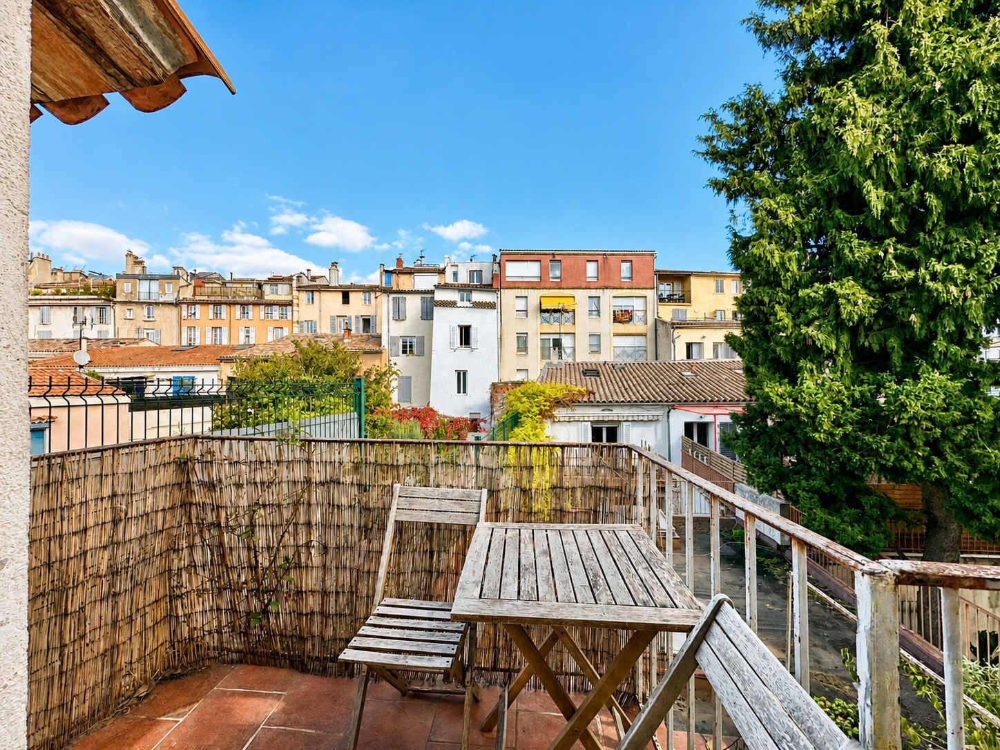 Un balcon avec une table et des chaises en bois, une clôture en bambou et une vue sur les bâtiments.