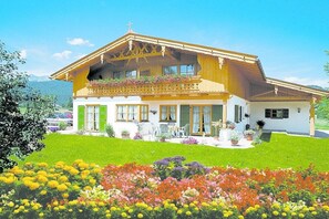 A two-story house with a sloped roof, a balcony with plants, and a well-maintained garden with various flowers. - Holiday apartment in Krün with swimming pool (Krün)