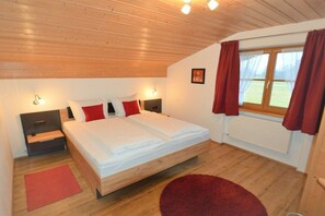 A bedroom with a wooden ceiling, a bed with white and red bedding, two bedside tables, a nightstand, a window with curtains, and a red rug on the floor. - Holiday apartment in Krün with swimming pool (Krün)