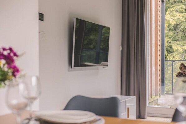 A room with a TV mounted on the wall, a dining table with chairs, and a window with curtains.