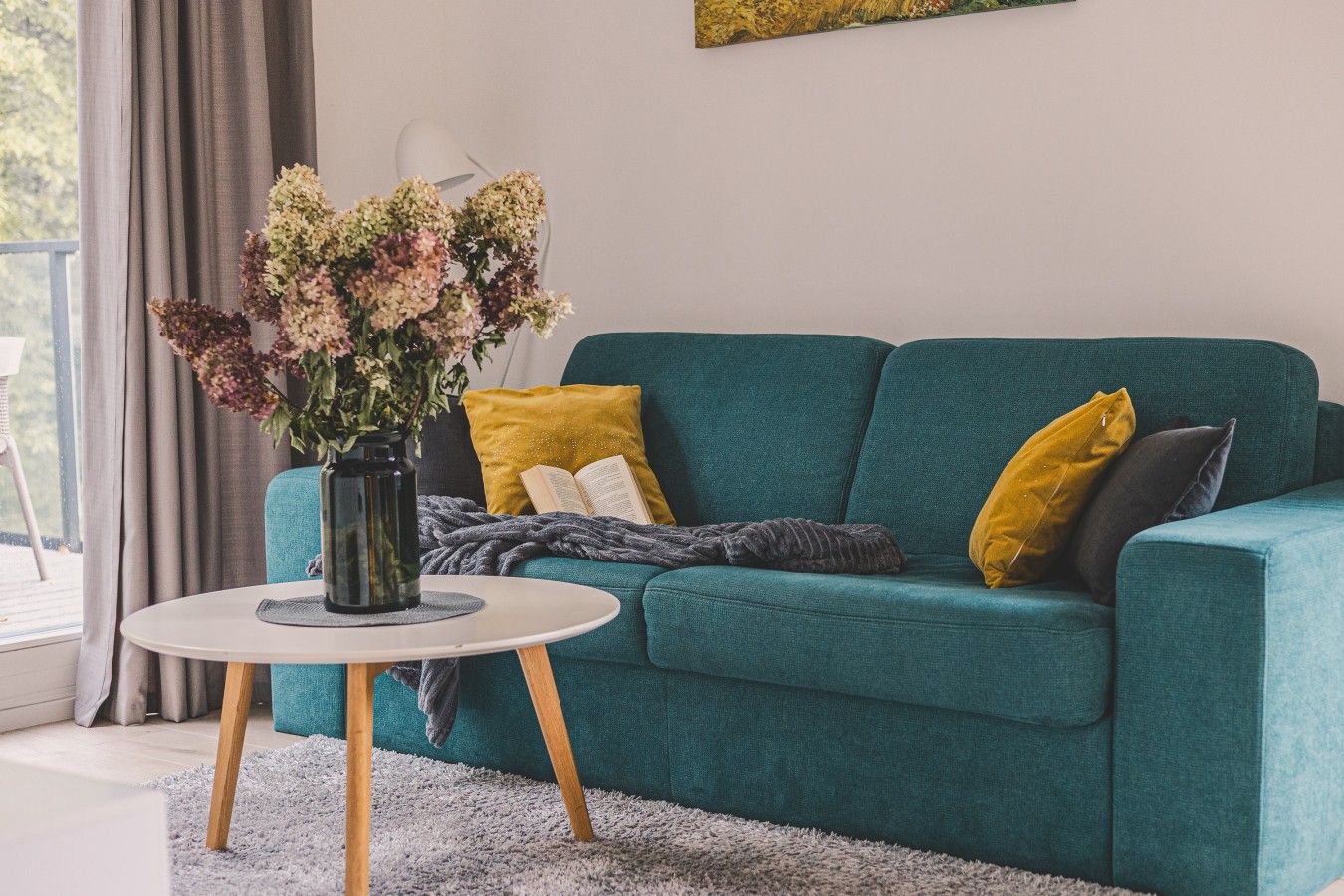 A teal sofa with yellow and gray cushions, a white round table with a vase of flowers, and a gray blanket on the floor.