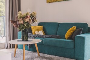 A teal sofa with yellow and gray cushions, a white round table with a vase of flowers, and a gray blanket on the floor.