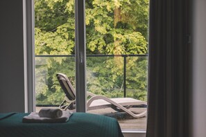 A room with a bed, a window with a view of greenery, and a chair outside.