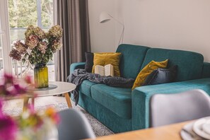 A teal sofa with cushions, a small round table, and a vase with flowers.