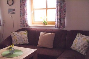 A living room with a sofa, a small table with a plant, a clock on the wall, and a window with curtains. - Holiday apartment with 1 bedroom (Fischbachau\/Birkenstein)