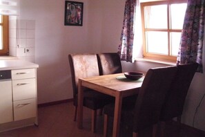 A dining area with a wooden table and chairs, a bowl on the table, and a window with curtains. - Holiday apartment with 1 bedroom (Fischbachau\/Birkenstein)