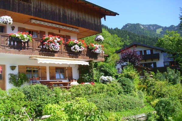 A traditional chalet with a balcony adorned with flower baskets, surrounded by lush greenery and a mountainous backdrop. - Holiday apartment with 1 bedroom (Fischbachau\/Birkenstein)