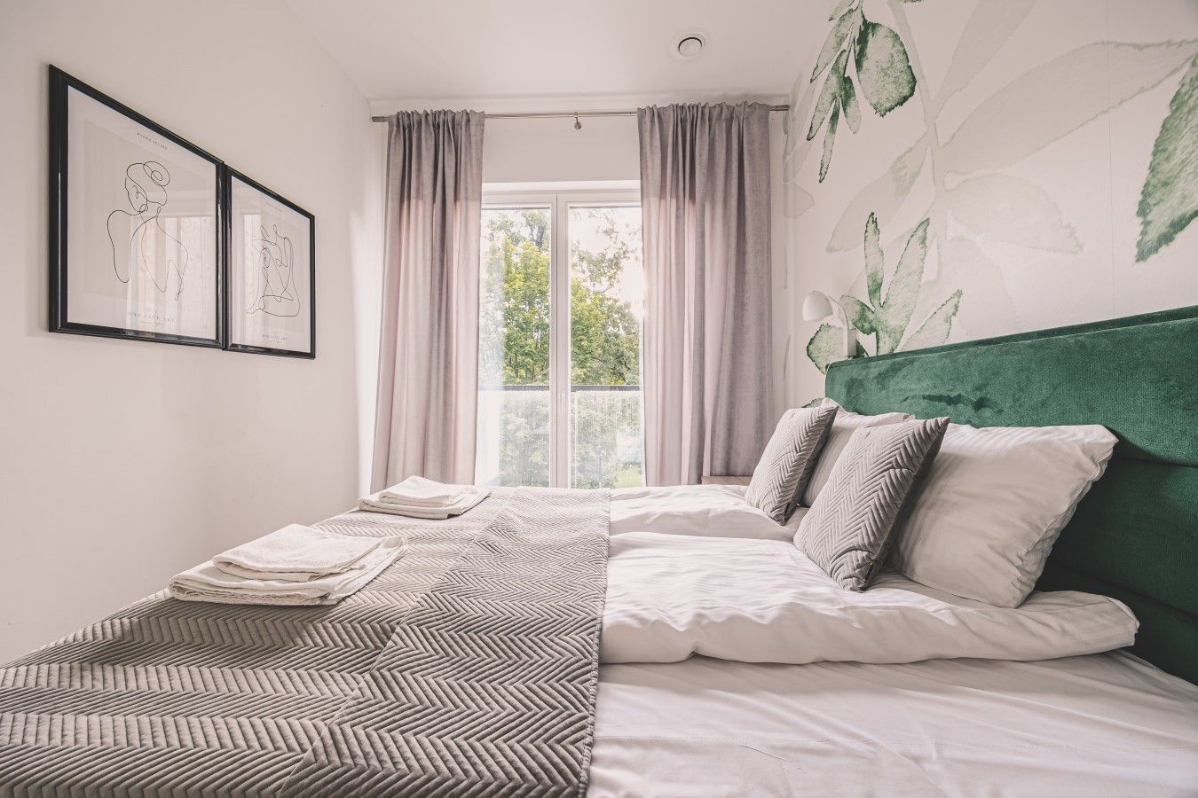 A bedroom with a bed, a green headboard, a window with curtains, and a wall with a leaf pattern.