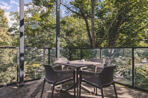 An outdoor seating area with a table and chairs, surrounded by a glass railing, overlooking a garden with trees.