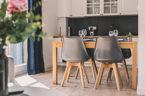 A dining area with a wooden table and two chairs, a kitchen in the background, and a window with curtains.