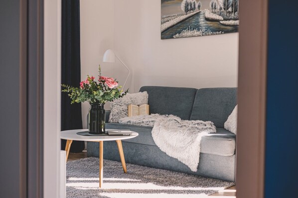 A living room with a grey sofa, a white round table with a vase of flowers, a fluffy throw blanket, and a painting on the wall.
