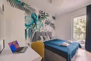 A bedroom with a bed, a desk with a laptop, a chair, and a large window.