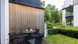 Une terrasse extérieure avec une clôture en bois, une table ronde entourée de chaises et un jardin agrémenté de haies et d’arbres.