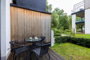An outdoor patio with a wooden fence, a round table with chairs, and a garden with hedges and trees. - Castor Apartament  | Z Tarasem przy Puszczy (Hajnówka)