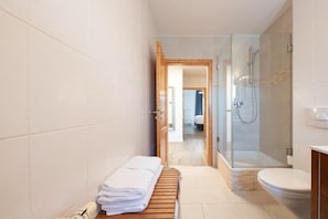 Casa de banho com chuveiro, sanita e um banco com toalhas.
