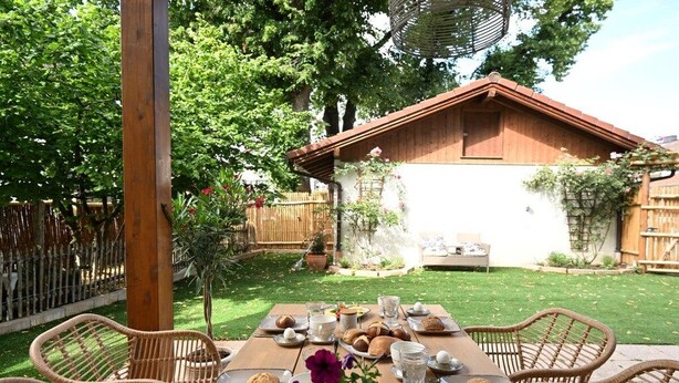Een terras met een houten tafel gedekt voor een maaltijd, omringd door rieten stoelen, met een huis en tuin op de achtergrond.