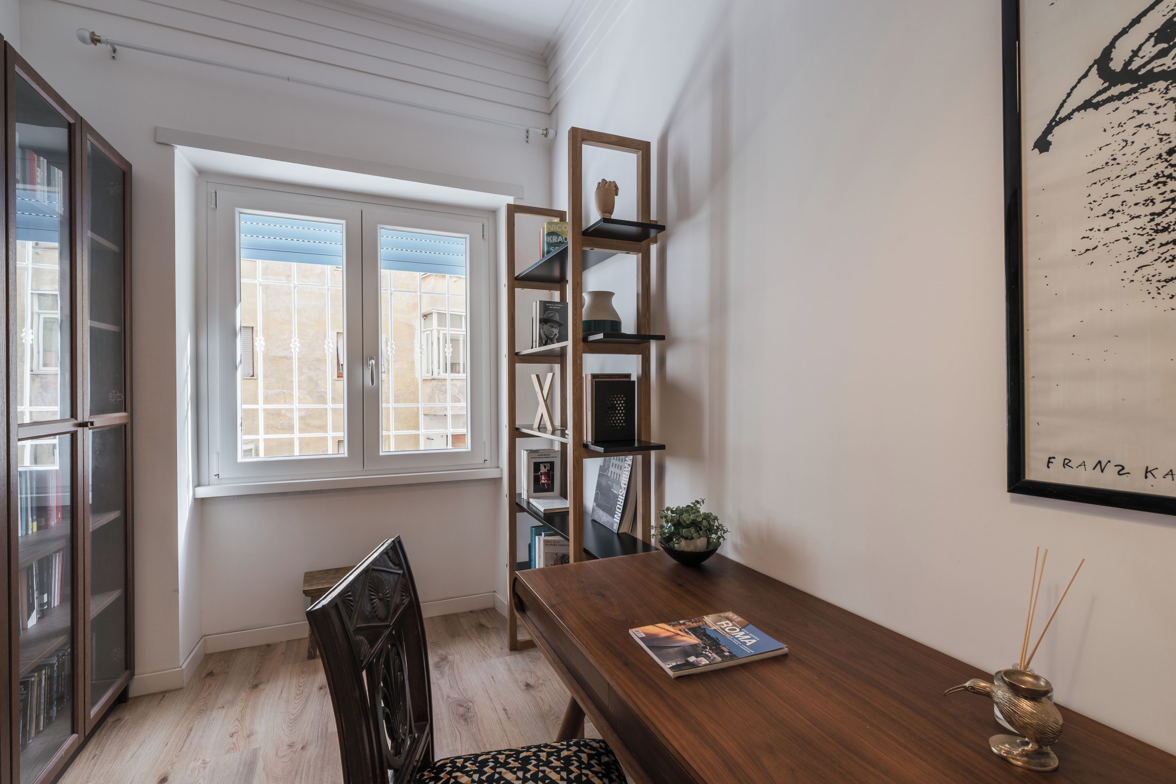 Ein Zimmer mit einem Holztisch, einem Bücherregal, einem Fenster mit Blick und einem gerahmten Kunstwerk an der Wand.