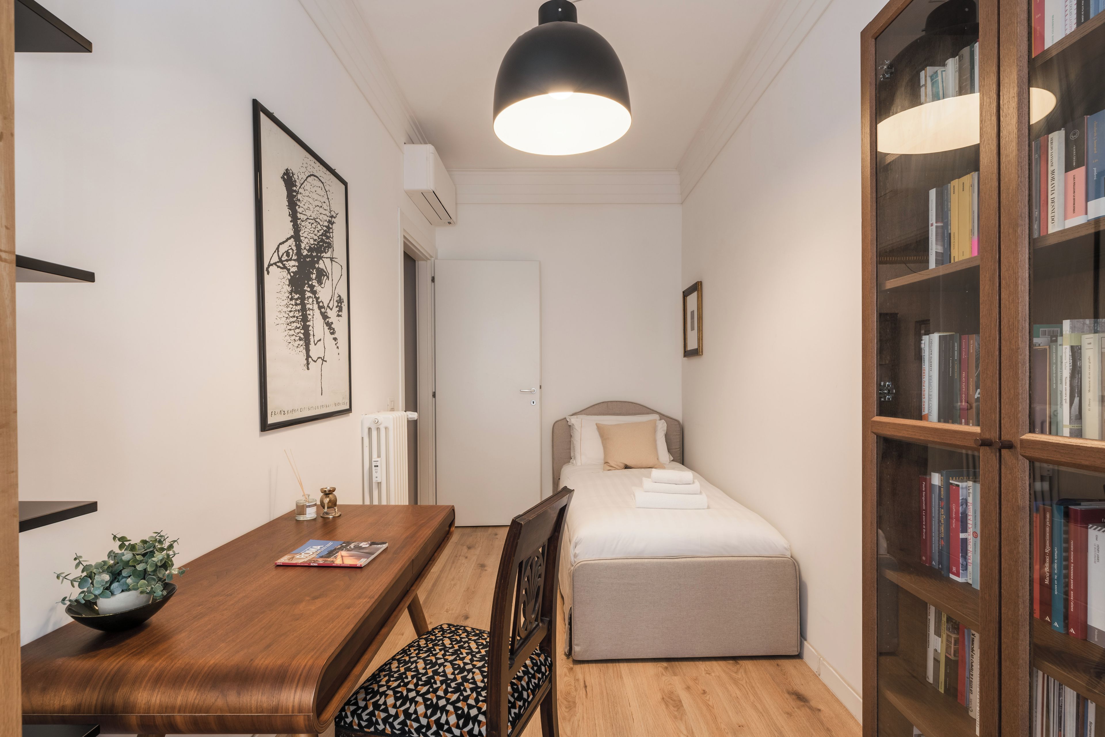 Ein gemütliches Schlafzimmer mit einem Einzelbett, einem Nachttisch mit Lampe, einem Bücherregal und einem gerahmten Kunstwerk an der Wand.
