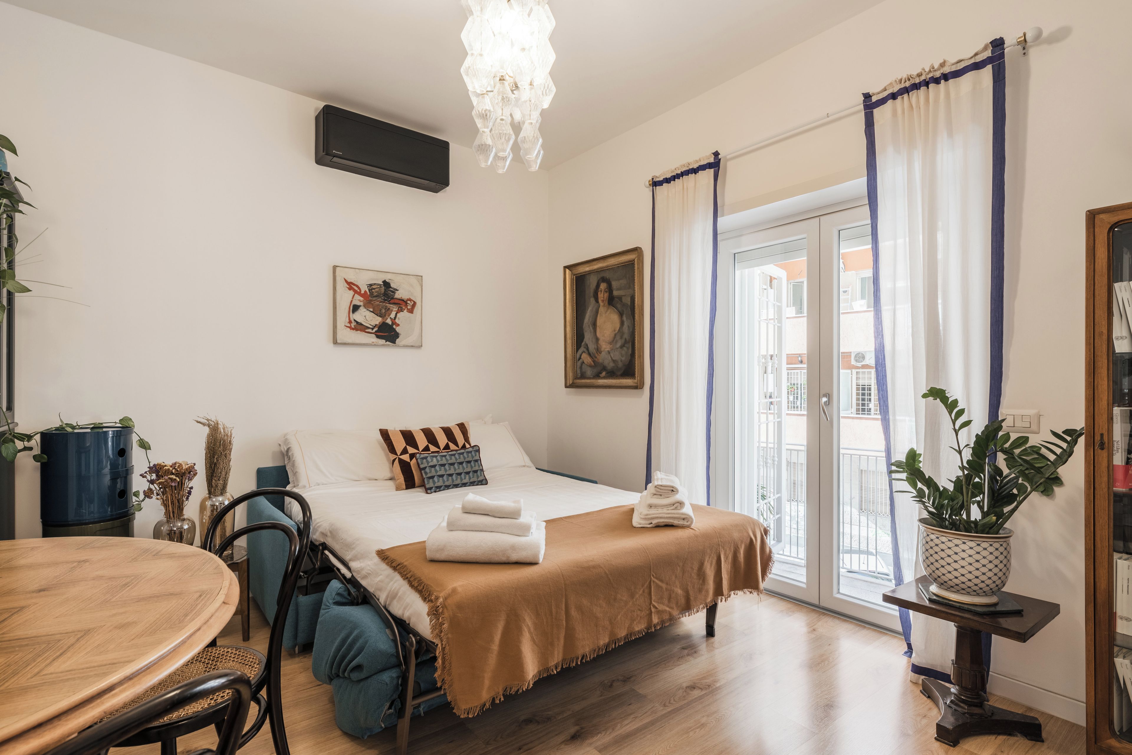 A hotel room with a bed, a dining table, a chair, a potted plant, and a painting on the wall.