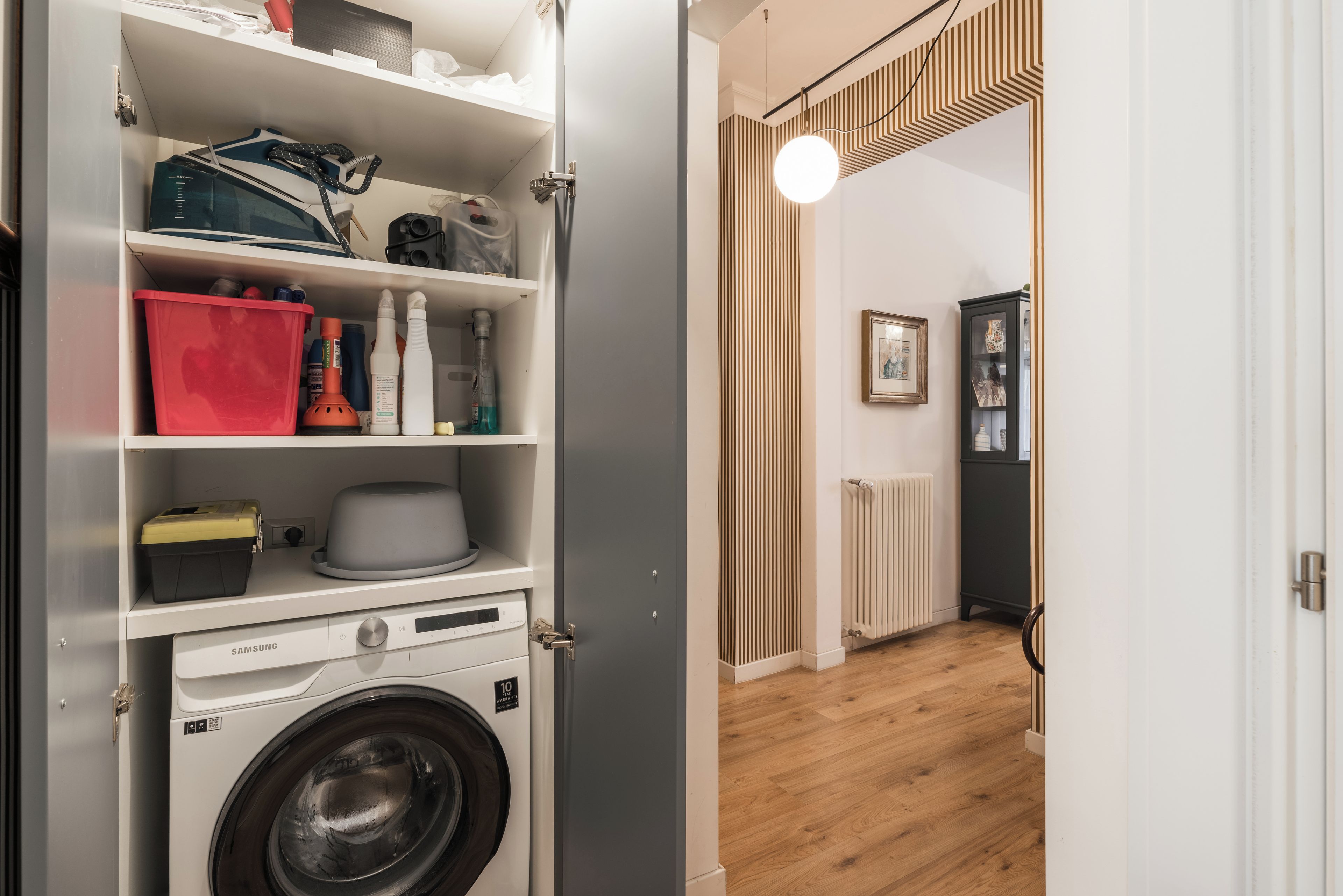 A laundry area with a washing machine, shelves with various items, and a hallway leading to a room with a radiator and framed pictures.
