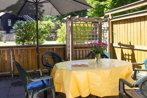Eine Terrasse im Freien mit einem runden Tisch, der mit einer gelben Tischdecke bedeckt ist, umgeben von Stühlen und einem Sonnenschirm, der Schatten spendet.