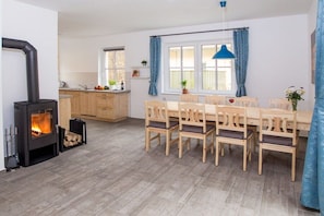 A modern kitchen with a wood-burning stove, a dining area with wooden chairs, and a window with blue curtains. - Ilsestein - Detached house (Ilsenburg)
