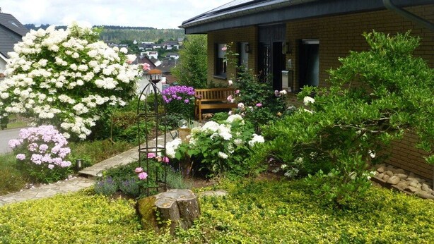 A residential building with a brick exterior, a wooden bench, and a garden with various plants and flowers. - "To the dike warden" (Losheim am See)