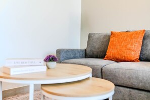 A living room with a grey sofa, an orange pillow, a small table with a white base, and a purple flower in a white pot. - HAVN - Studio in Regalia, Business Bay (Dubai)