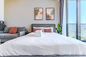 A hotel room with a large bed, a grey sofa, two framed wall art pieces, and a view of the city through the window. - HAVN - Studio in Regalia, Business Bay (Dubai)