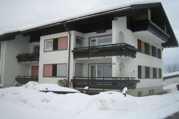 Edifício de vários andares com varandas, entrada coberta e neve acumulada ao redor da propriedade.