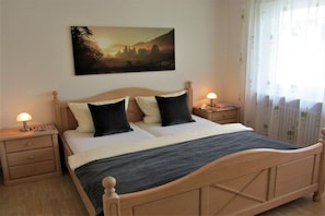 Quarto com cama de madeira, duas mesas de cabeceira, um criado-mudo, um quadro na parede e uma janela com cortinas.