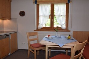Uma cozinha pequena com uma mesa de jantar posta para dois, um relógio na parede e uma janela com cortinas.