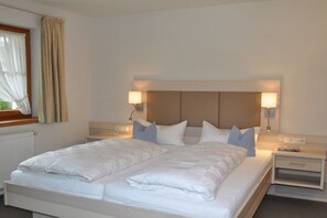 Cama de casal com roupa de cama branca, duas luminárias de cabeceira, mesa de cabeceira e janela com cortinas.