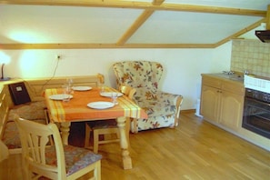 A dining area with a wooden table, chairs, and a floral armchair. A kitchen with wooden cabinets and an oven is visible in the background.