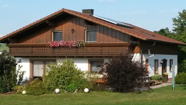 Uma casa de madeira de dois andares, com telhado inclinado, varanda frontal e gramado bem cuidado.