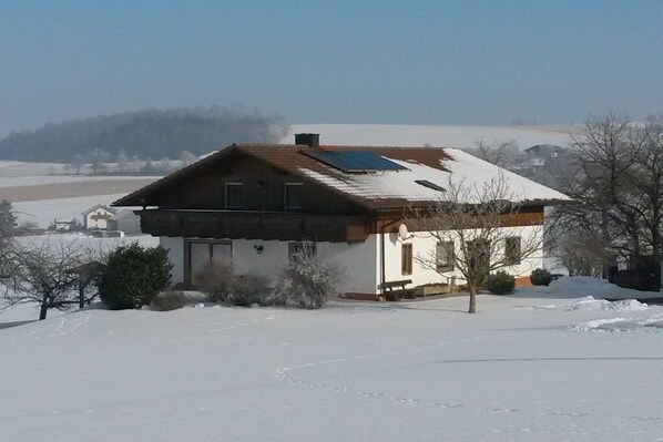 A house with a sloped roof, surrounded by snow-covered ground, with a clear sky in the background.