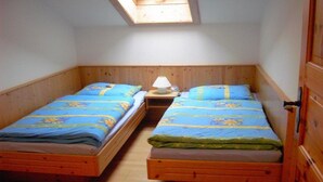 Quarto pequeno com duas camas de madeira, cada uma com colcha estampada em azul e amarelo, uma mesinha de cabeceira com luminária e um claraboia acima.