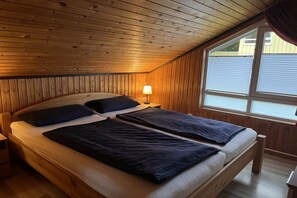 A room with a sloped ceiling, wooden walls, a bed with two pillows, a bedside lamp, and a window with blinds. - Holiday home in Extertal-Rott with barbecue (Extertal-Rott)