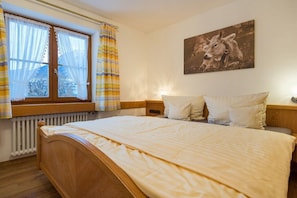 Quarto com cama de madeira, janela com cortinas, quadro de vaca na parede e mesa de cabeceira com luminária.
