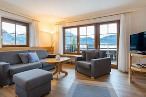 Uma sala de estar moderna com sofá, banquinho e poltrona, uma mesinha redonda, televisão e grandes janelas com vista para as montanhas.