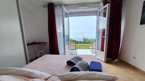 Een slaapkamer met een bed, kussens, een blauwe handdoek en uitzicht op een balkon met stoelen.