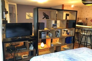 Ein Zimmer mit einem Bett, einem Fernseher, einer Holzhockerreih mit verschiedenen Gegenständen sowie einer Küchenzeile mit Herd und Stühlen.
