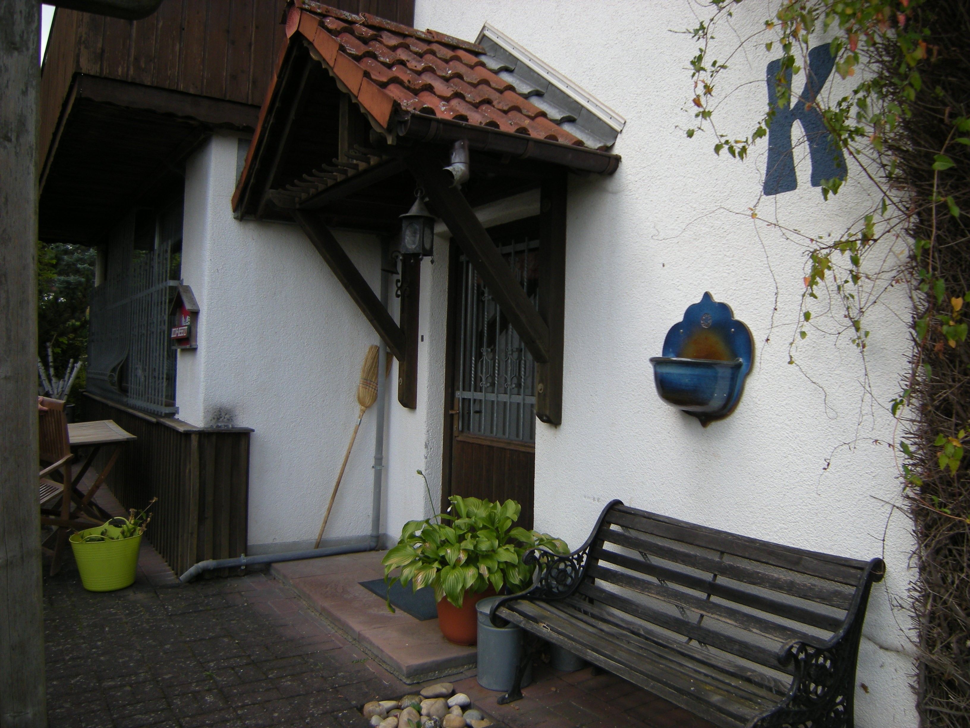 A building with a white exterior, a wooden bench, a potted plant, and a wall-mounted light fixture.