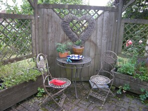 Um pátio de jardim com treliça em formato de coração, duas cadeiras de metal, uma mesinha com uma tigela e plantas em vasos.
