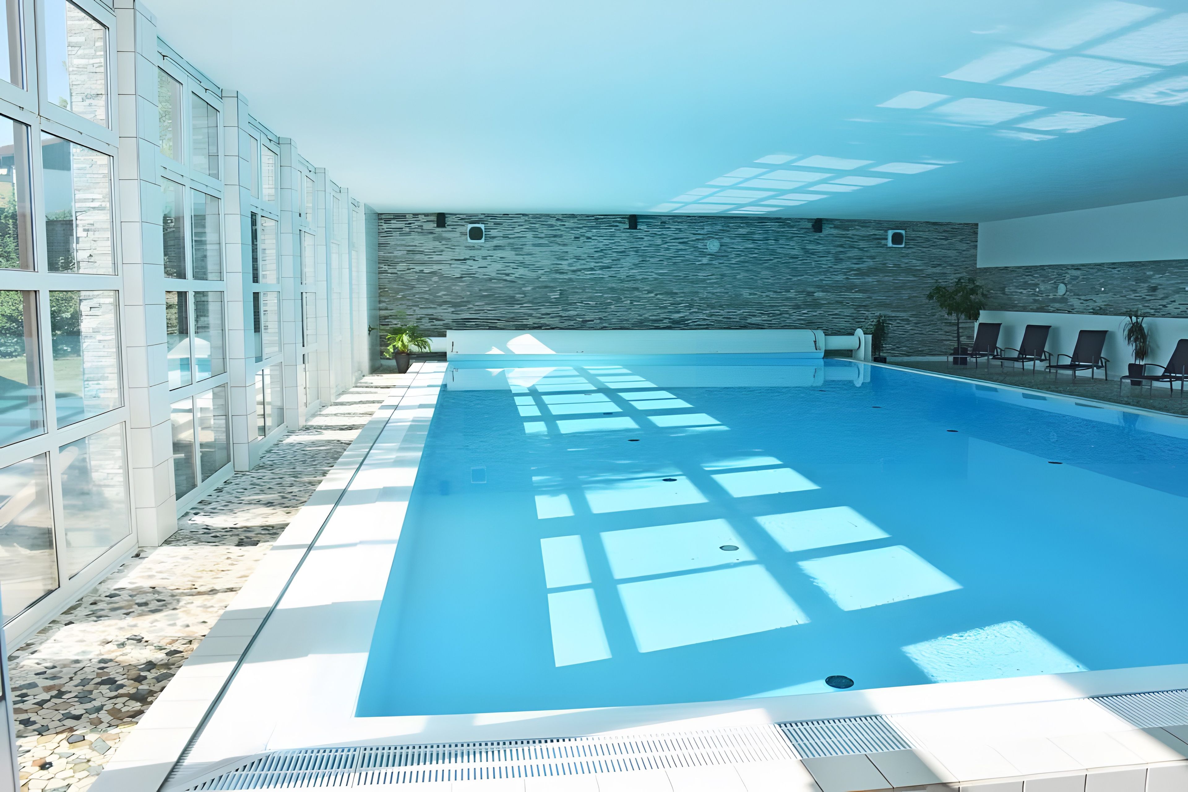 Ein moderner, überdachter Schwimmbereich mit großen Glaswänden, einer Steinwand und einer von der Sonne durchfluteten Decke.