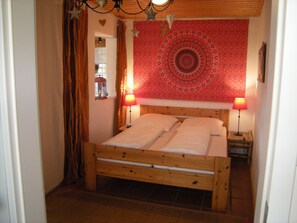 Uma cama de madeira com duas almofadas, um tapeçário vermelho estampado na parede e duas luminárias de cabeceira.