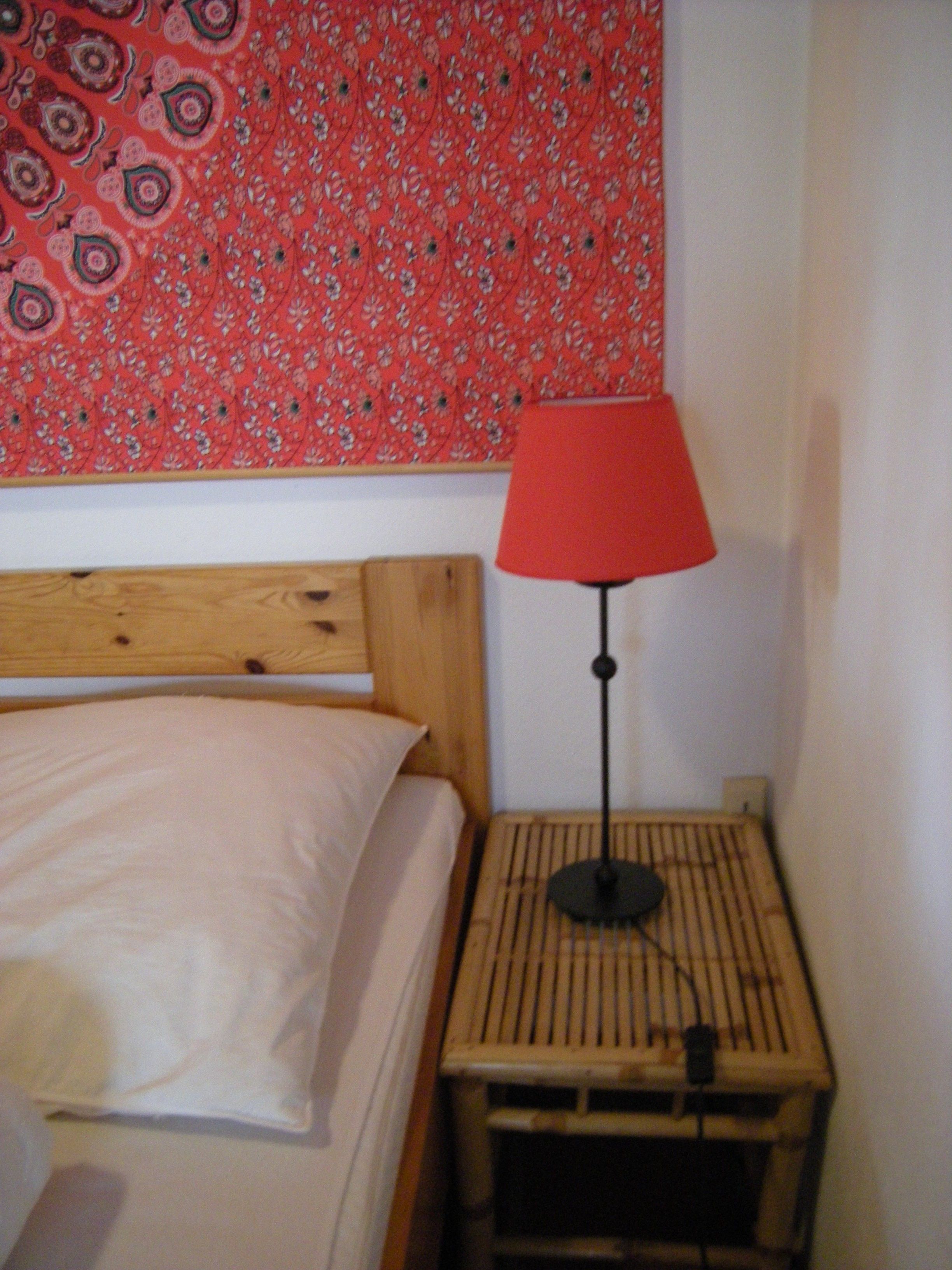A bed with a wooden headboard, a red lamp on a bedside table, and a tapestry with a floral pattern on the wall.