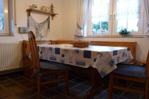 Ein Essbereich mit einem Tisch, der mit einer gemusterten Tischdecke bedeckt ist, Holzstühle, ein Korb auf dem Tisch und ein Fenster mit Vorhängen.