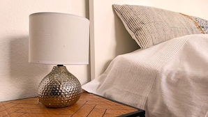 Ein Nachttisch mit Lampe und einer strukturierten Vase, ein Bett mit weißer Bettdecke und einem gemusterten Kissen.